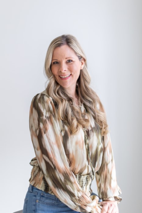 A professional headshot of Cruisinjules, a blonde woman with her hair curled sitting in front of a gray background.